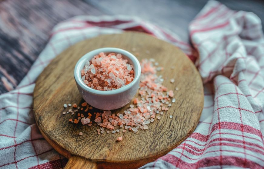 Small white ceramic bowl of pink Himalayan salt on a round wood platter with a white and red textile in the background