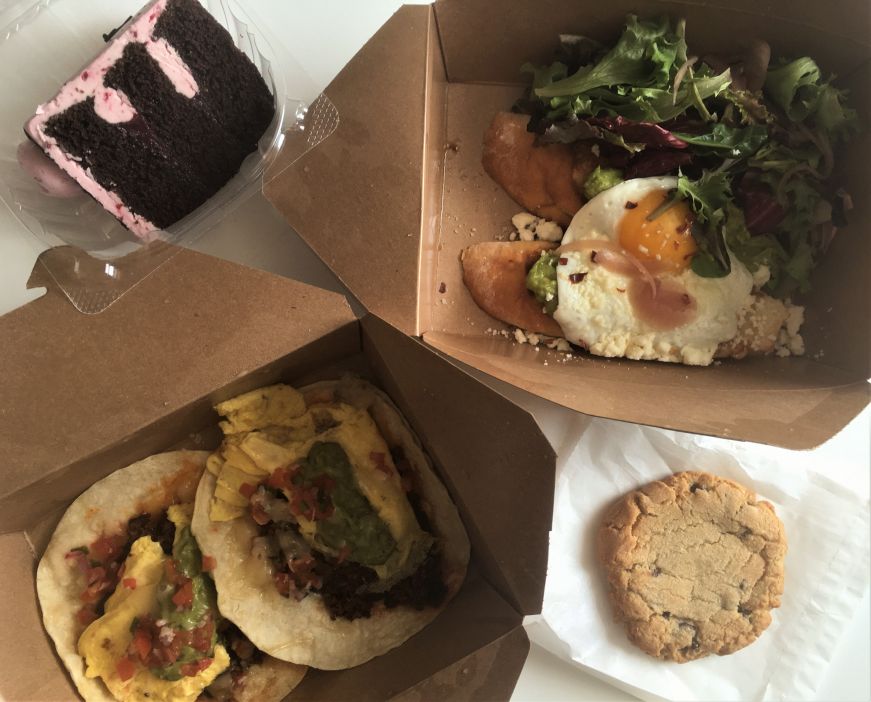 Takeout containers with raspberry chocolate cake, avocado toast, chocolate chip cookie, and breakfast tacos