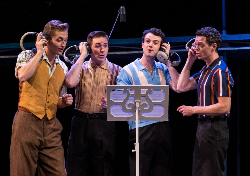 Four men in 1960s-era clothing singing into a microphone