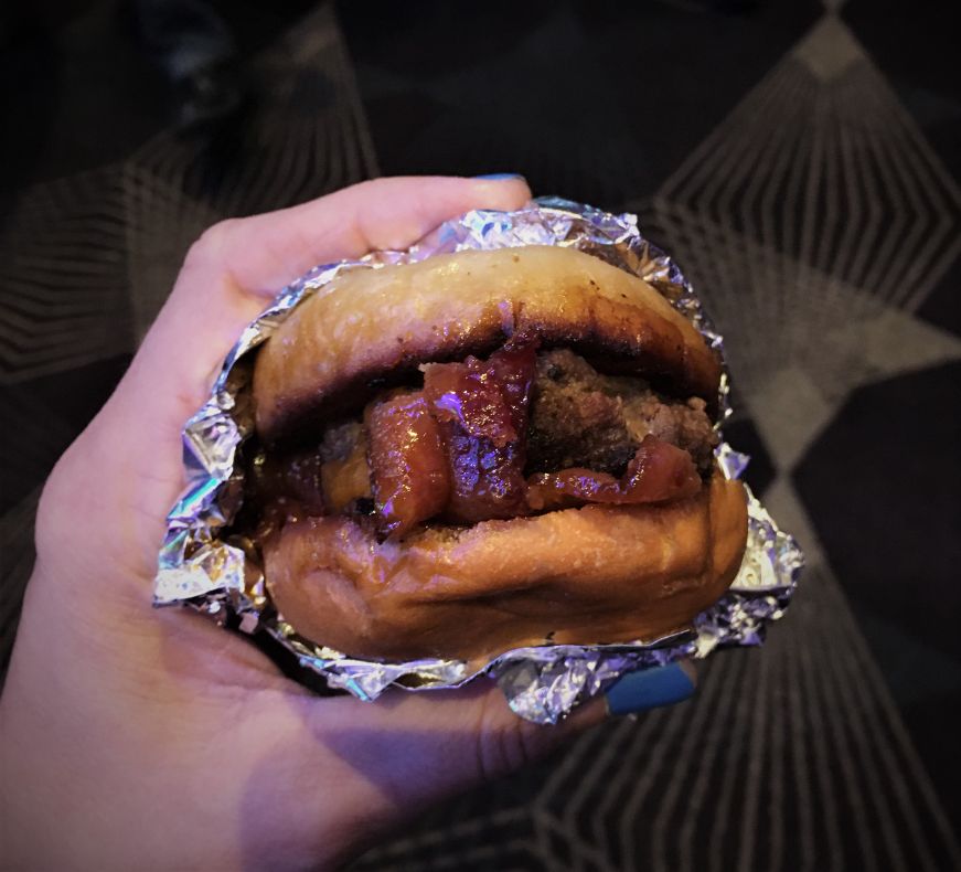 Hand holding a small bacon cheeseburger partially wrapped in foil