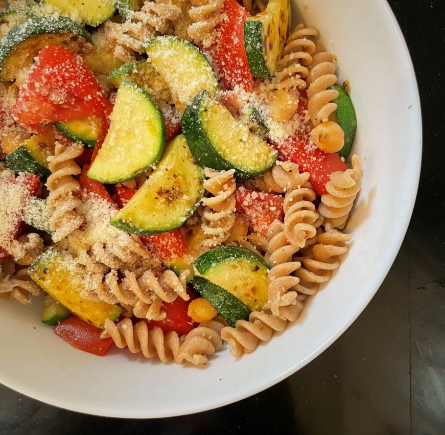 White bowl filled with rotini pasta mixed with sauteed zucchini and tomato chunks sprinkled with grated Parmesan