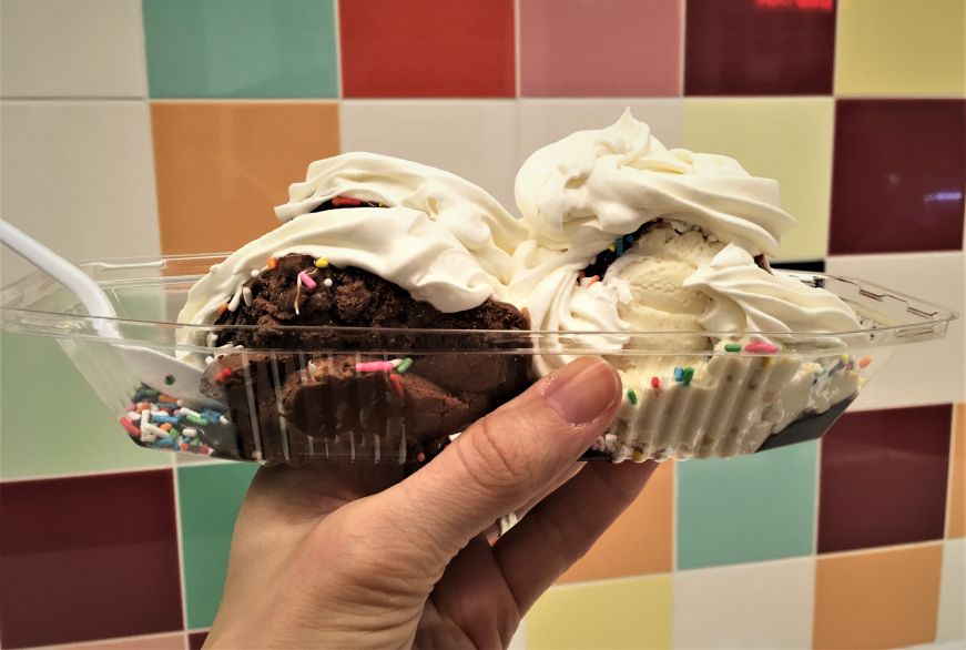 Hand holding large ice cream sundae with whipped cream and sprinkles against a multi-colored tile wall