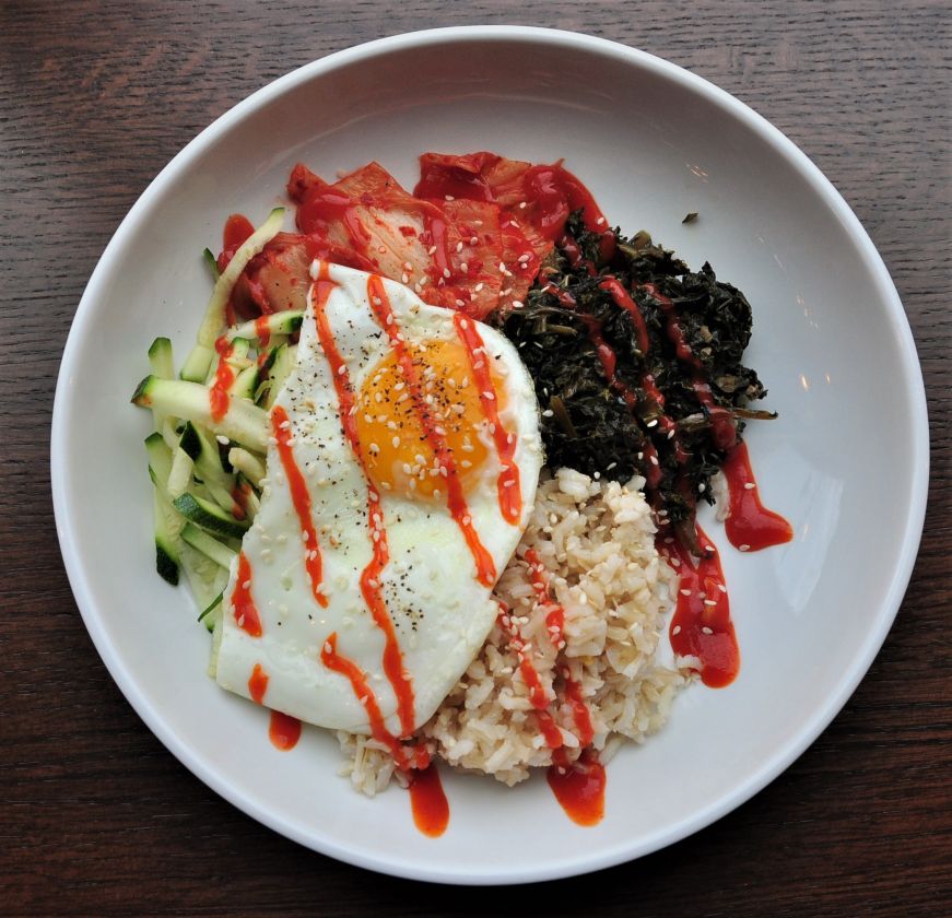 Plate of rice, fried egg, kimchi, and kale drizzled with hot sauce