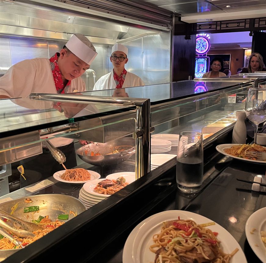 Two chefs preparing noodle dishes and diners sitting a counter looking on