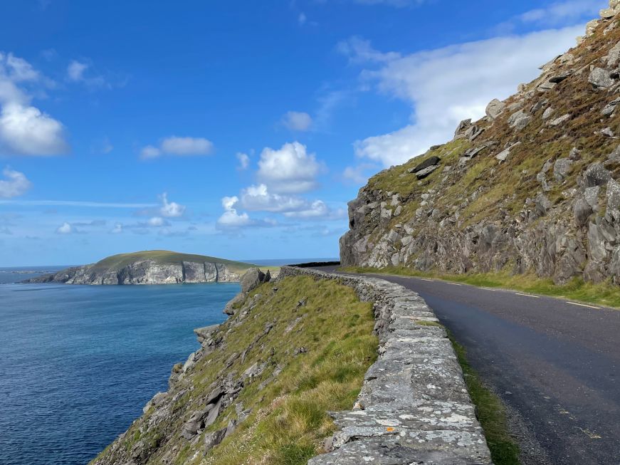 Blacktop road running along the ocean with a sheer drop on one side and a rock wall on the other