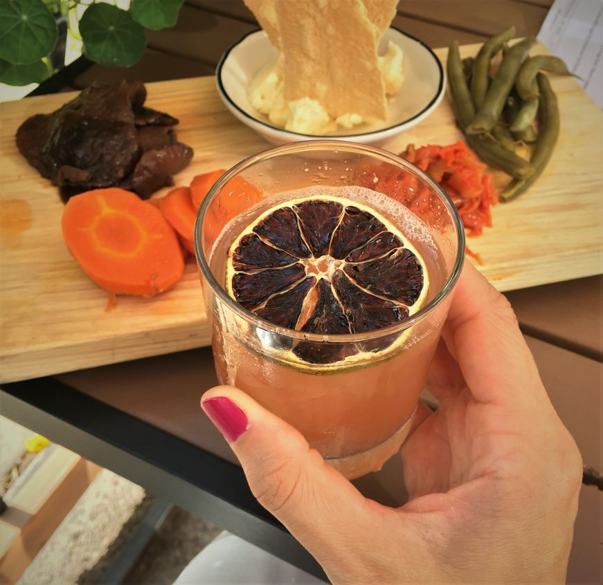 Hand holding a cocktail garnished with a dried orange slice with a board of pickled vegetables in the background