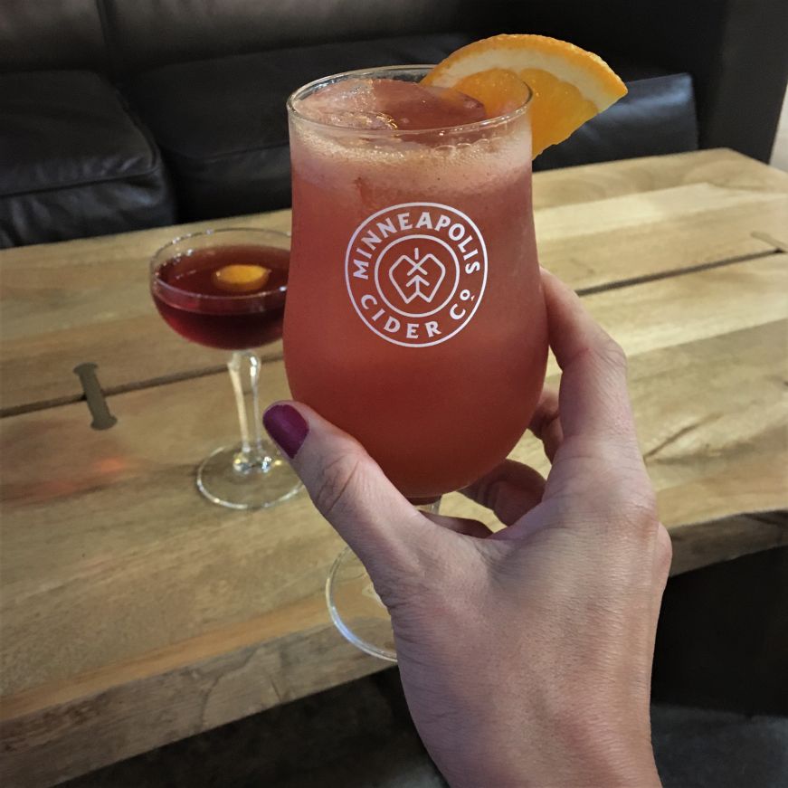 Spirit of the Toucan cocktail, Minneapolis Cider Co. Hand holding a pink cocktail garnished with an orange slice