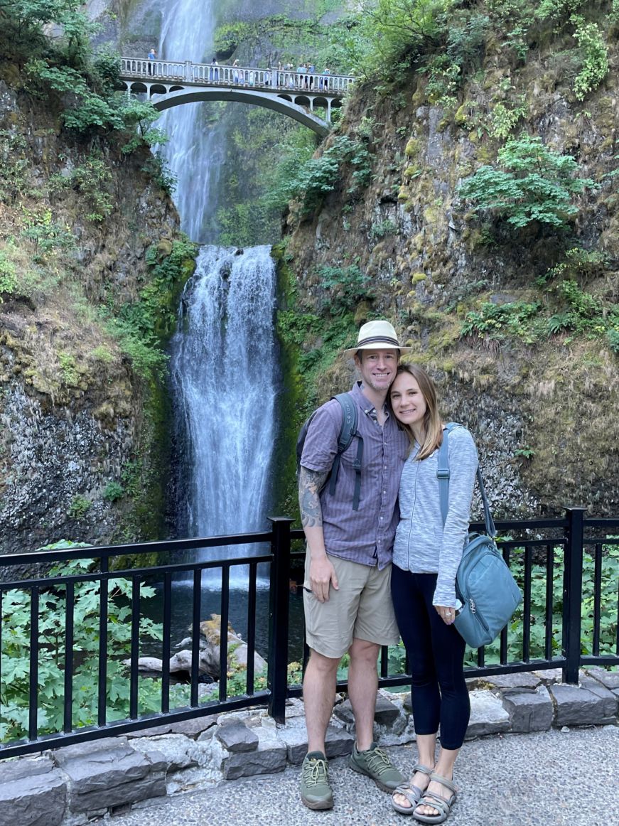 Josh and Stacy standing in front of a tall waterfall with a bridge in the background