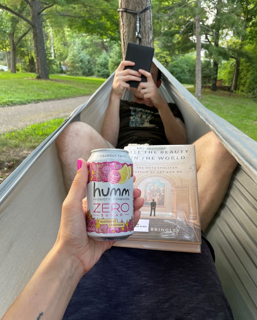 Stacy's legs in a hammock with Mike facing her.  Stacy is holding a can of kombucha and book, and Mike is reading an e-reader