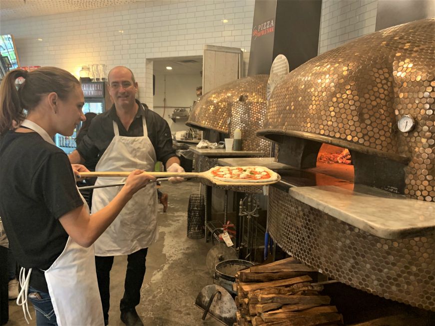 Stacy putting pizza in oven with pizza peel while chef looks on, 1889 Pizza Napoletana, Kansas City, Kansas