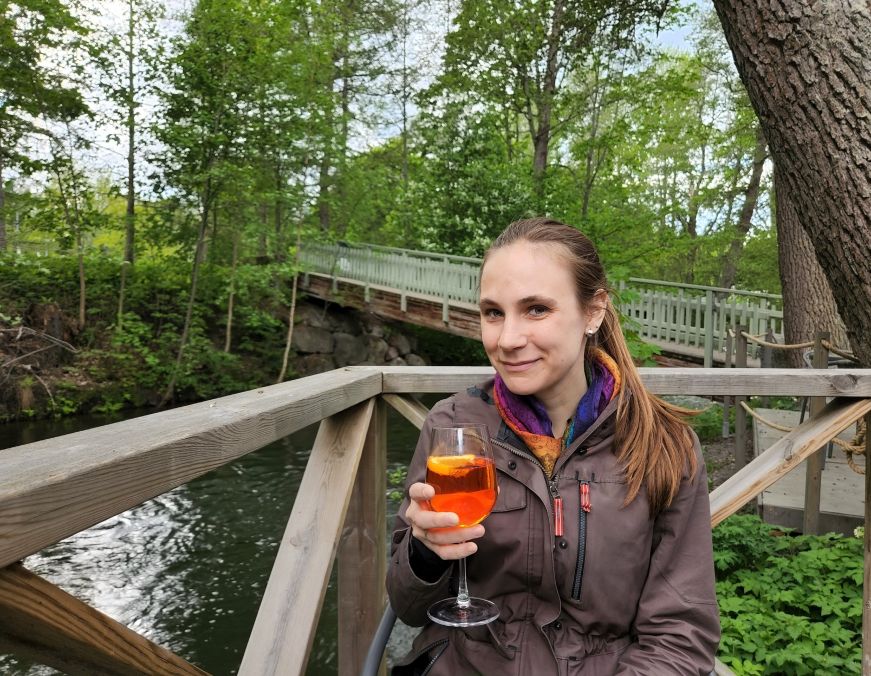 Stacy with an Aperol spritz at Wine&Cafe Jokiranta Stacy holding an Aperol spritz with a river and trees in the background