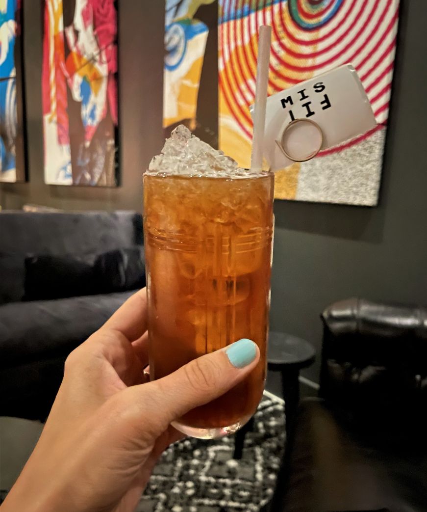 Sunglasses at Night, Stilheart Distillery & Cocktail Lounge, Minneapolis Stacy's hand holding a coffee-colored cocktail in a highball glass with colorful modern art in the background