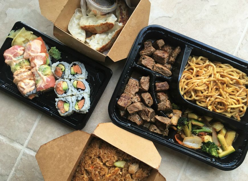 Takeout containers with sushi rolls, dumplings, fried rice, and pieces of steak with vegetables and noodles
