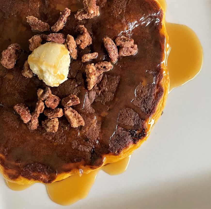Close up view of sweet potato pancake topped with a dollop of whipped butter, maple syrup, and candied pecans