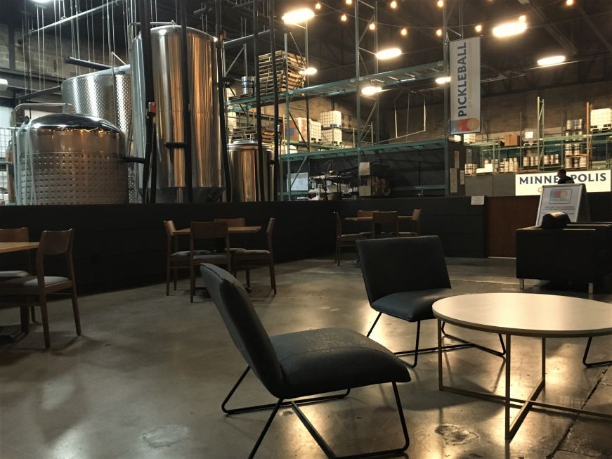 Taproom furnished with tables and chairs with brewing equipment in the background