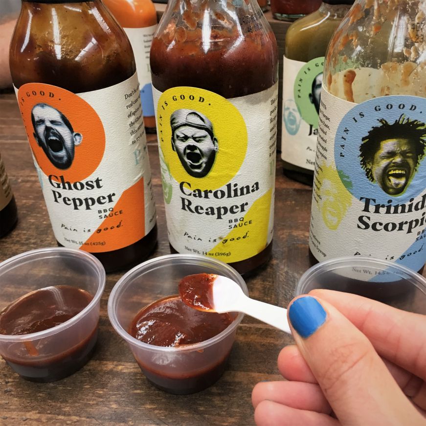 Hand holding small plastic spoon with barbecue sauce and bottles of barbecue in the background, Spicin Foods, Kansas City, Kansas