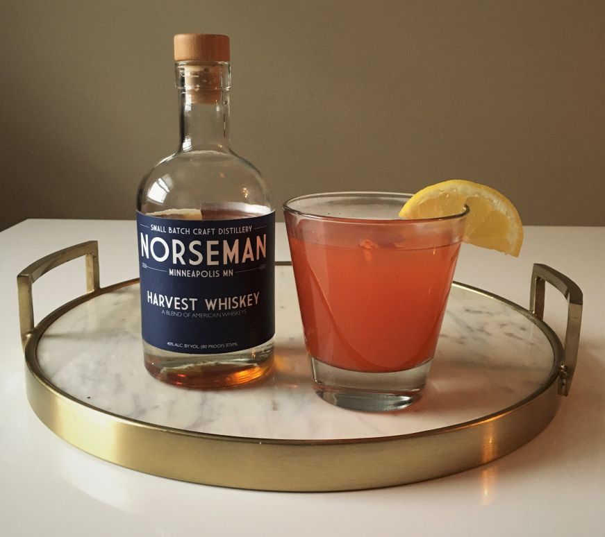 Bottle of Norseman Distillery whiskey on a marble tray with a pink cocktail garnished with a lemon slice