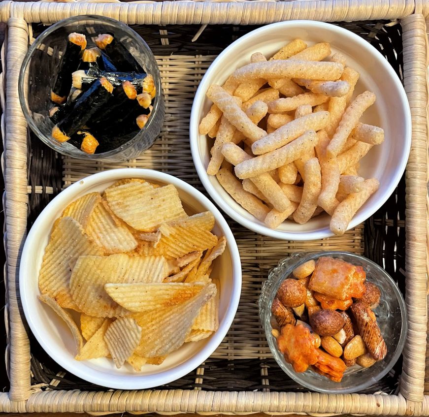 Four bowls of chips and rice crackers on a wicker tray