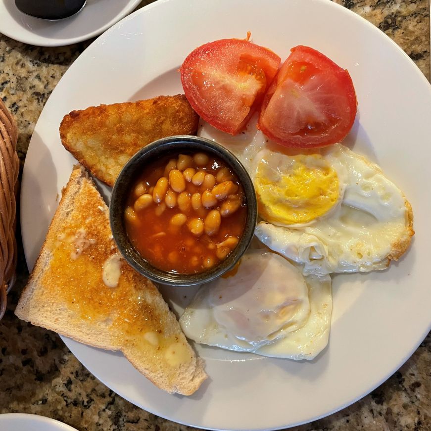 White plate with toast, fried eggs, tomatoes, and a cup of baked beans