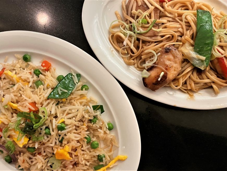 Top down view of a plate of vegetable fried rice and plate of lo mein noodles with chicken and vegetables