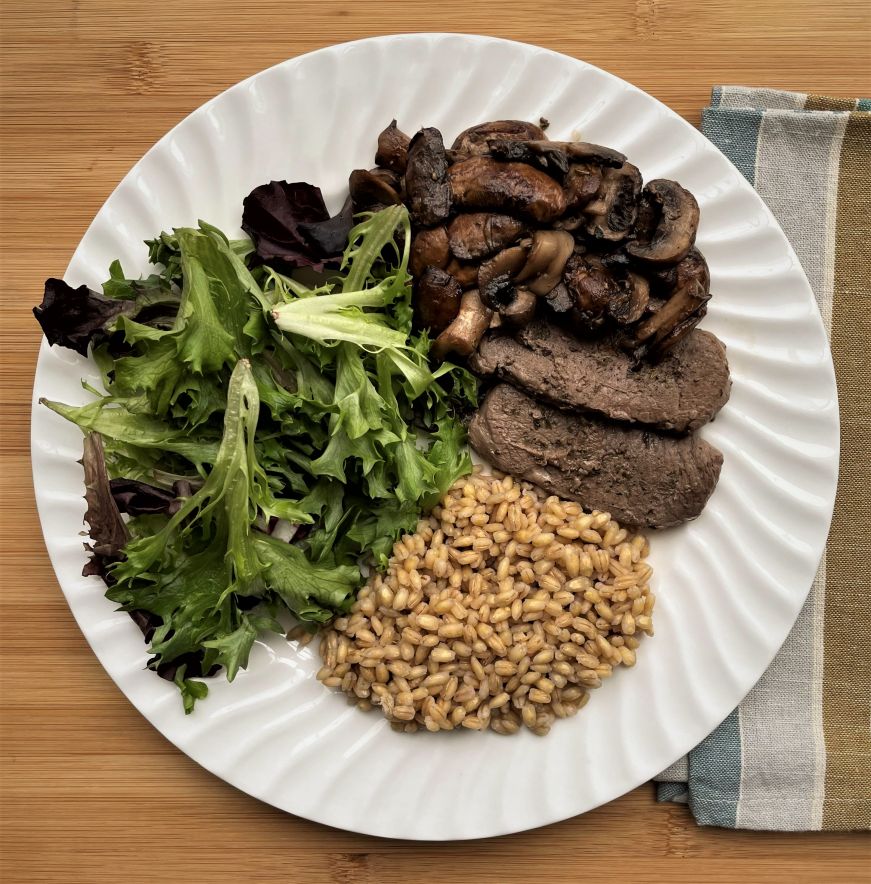 White plate with mixed greens, barley, vension tenderloin, and mushrooms