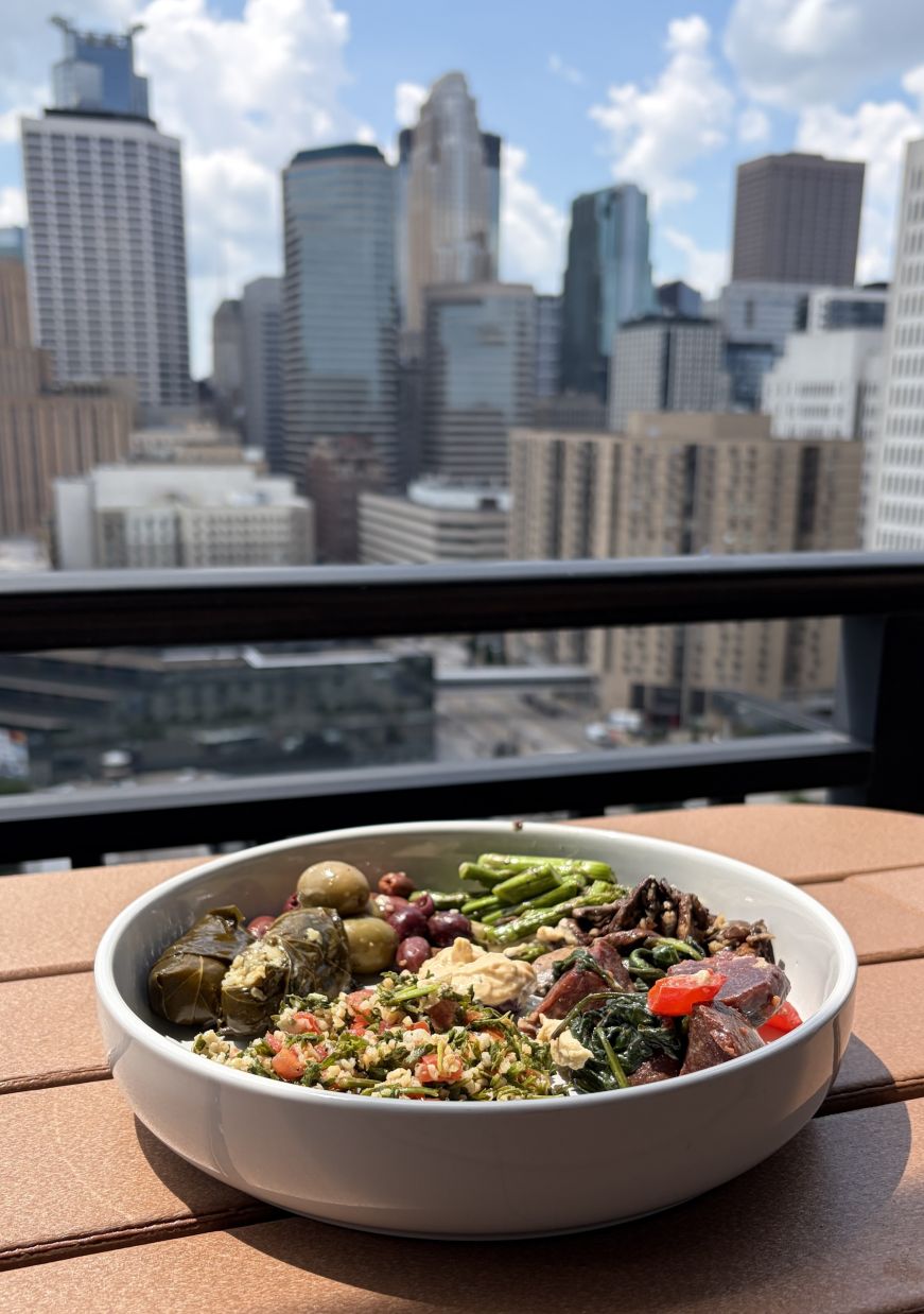 Bowl of tabbouleh, grape leaves, hummus, and vegetables with a skyline in the background