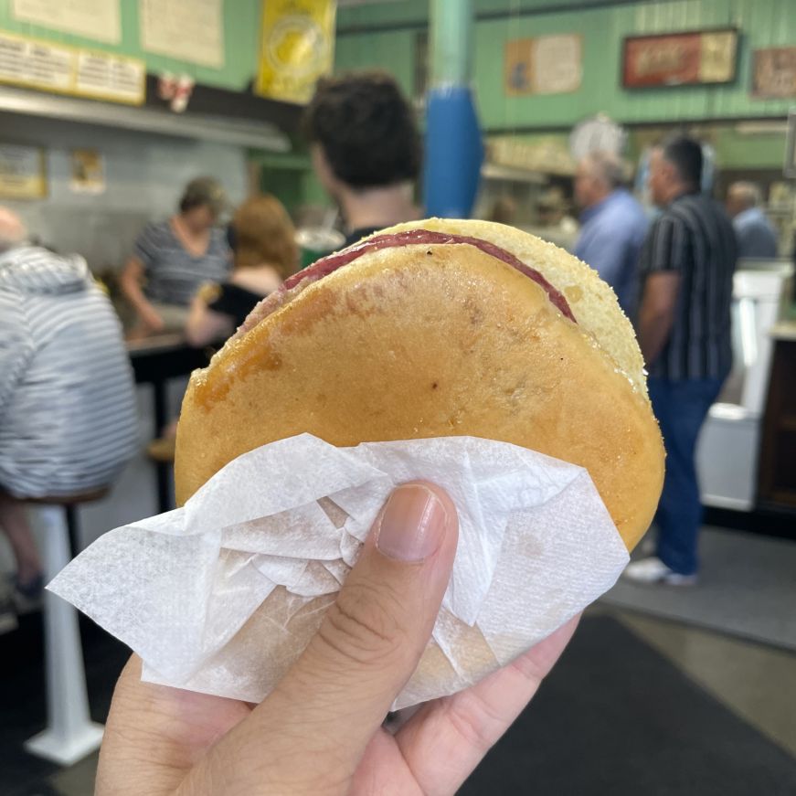 Stacy's hand holding a bologna and salami sandwich on a round roll