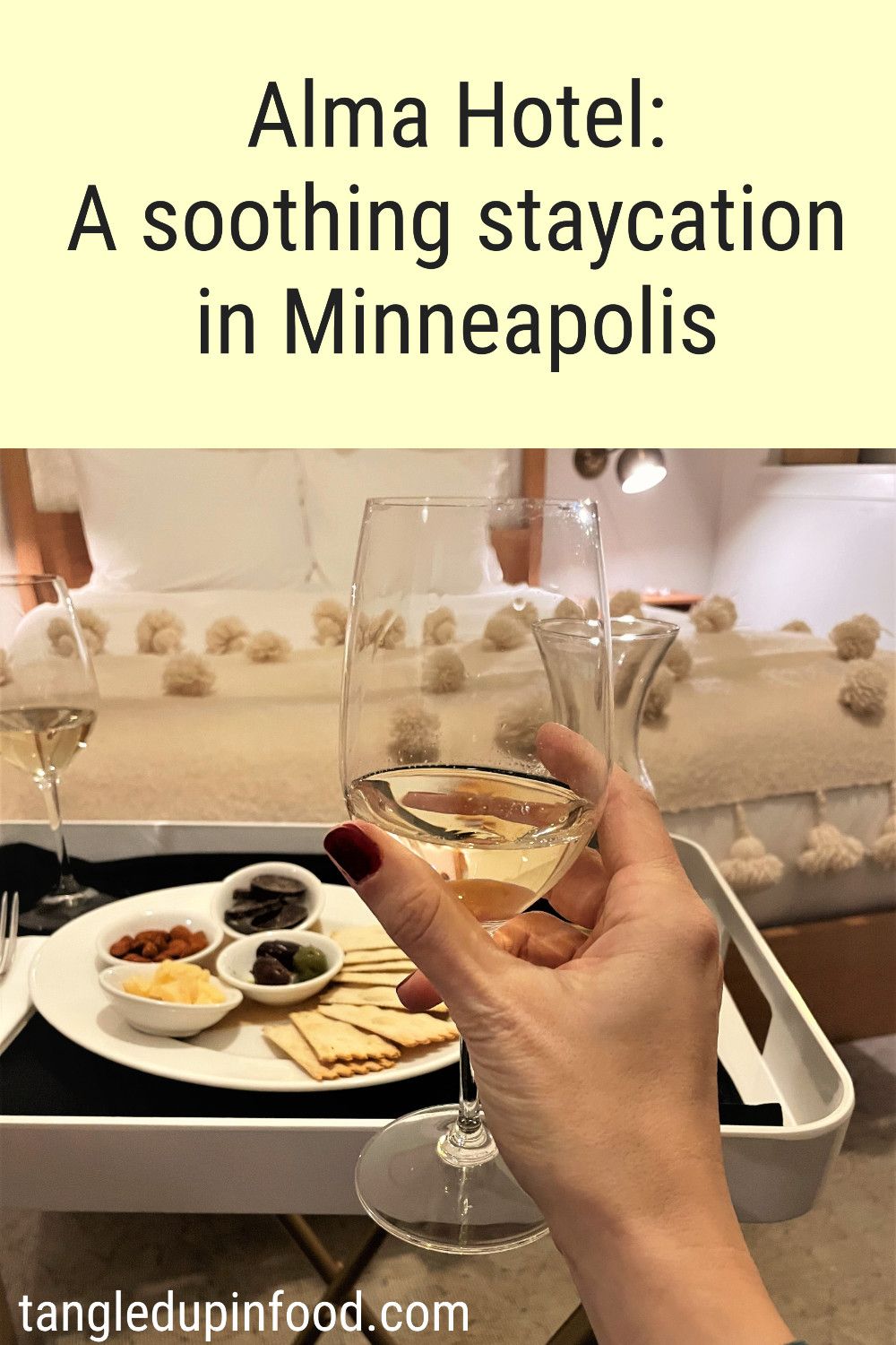Hand holding a glass of white wine with a tray of antipasti and a hotel bed in the background with text reading "Alma Hotel: A soothing staycation in Minneapolis"