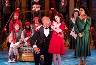 Red headed girl being embraced by a man in a tuxedo, surrounded the rest of cast in Christmas-themed costumes