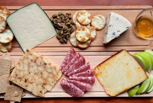 Two rectangular cheese boards with four blocks of cheese, sliced apples, salami, pistaschios, crackers, and a small bowl of honey