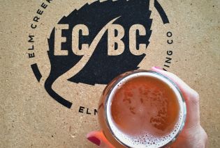 Top down view of hand holding beer over a table with the logo for Elm Creek Brewing Co.